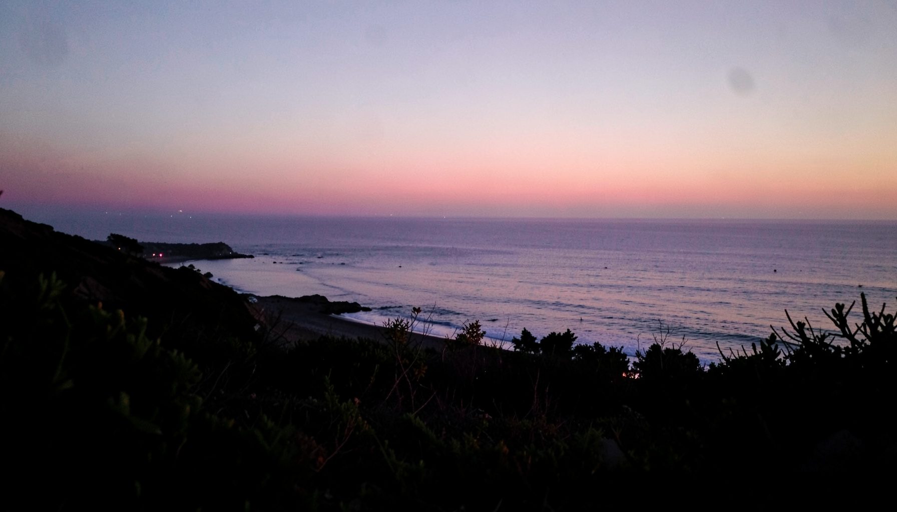 Paysage côtier au crépuscule avec une mer calme, un ciel aux tons pastel et une végétation de bord de mer en premier plan, évoquant la sérénité du voyage